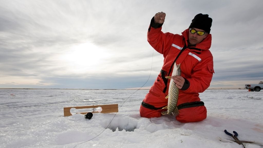 Bilden föreställer en man, iförd en röd flytoverall, på en is. Han håller på och isfiskar och har precis dragit upp en stor fisk.