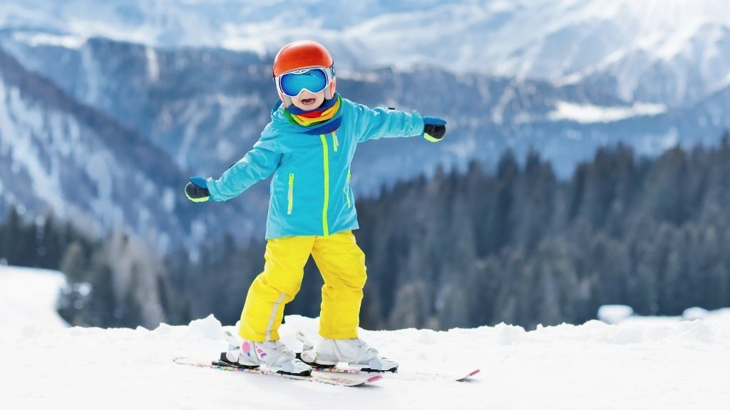 Bilden föreställer ett barn som står på skidor uppe på ett fjäll. Barnet har en turkos jacka, röd hjälm och gula termobyxor.