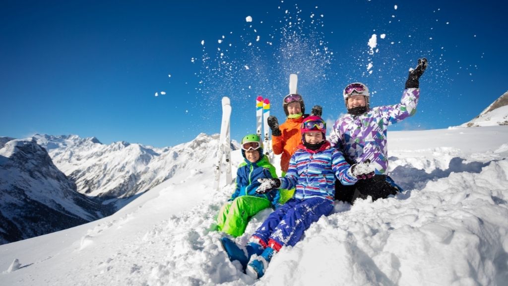 Bilden föreställer fyra barn som sitter uppe på ett snötäckt fjäll. Barna har skidkläder på sig och det står skidor bakom dem. Barna kastar upp snö ovanför sina huvud.
