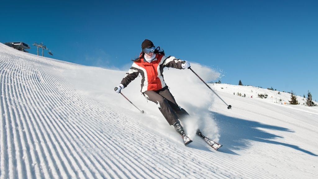 Bilden föreställer en kvinna som i hög fart åker ned för en backe med skidor. Hon är klädd i mörkgrå skidbyxor och hennes jacka är röd, vit och mörkgrå. Hon bär också hjälm och skidglasögon.