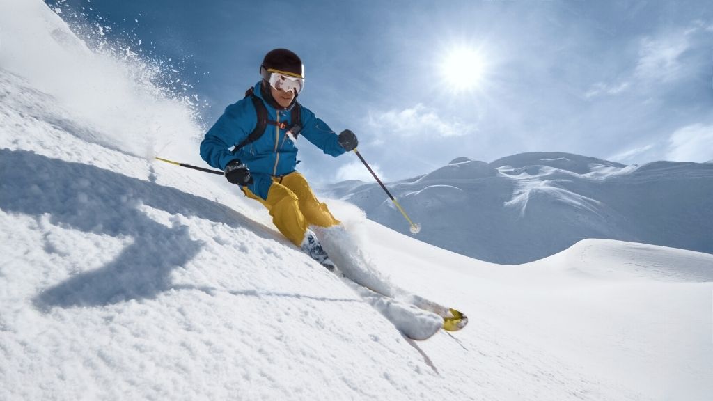 Bilden föreställer en man som åker skidor ner för ett snötäckt fjäll. Mannen har blå skidjacka, gula skidbyxor, svart skidhjälm och skidglasögon på sig. I bakgrunden lyser solen.